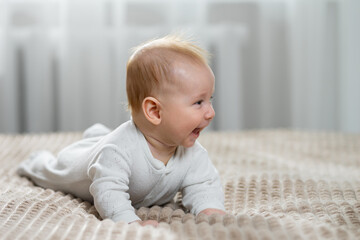 Fototapeta premium Happy baby dressed in white bodysuit lying on stomach and looking to the side, expressing joy and laughter during childhood development on a comfortable bed indoors