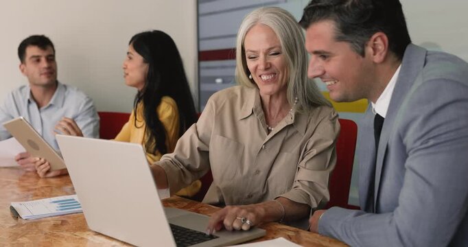 Happy mature business leader woman presenting online product, web project to positive male colleague, showing content on laptop at co-working office workplace, pointing at screen