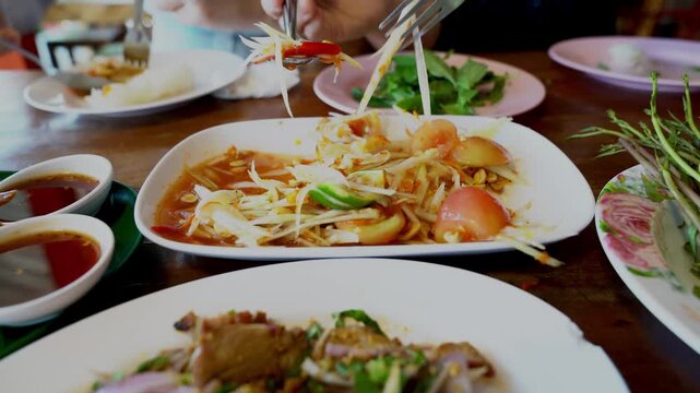 Thai spicy green papaya salad or Som Tum served on a white plate in a restaurant. Accompanied by various side dishes like spicy minced meat and fresh vegetables for an authentic dining experience.