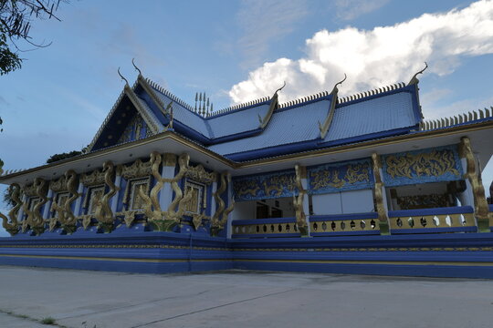 Exterior view of a blue Buddhist temple decorated with golden naga sculptures and traditional Thai architectural details under dramatic evening clouds in Mukdahan Province, northeastern Thailand.