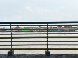 Fototapeta premium Yellow metal railing on a waterfront promenade looking over a wide river and a traditional riverside village under a cloudy sky