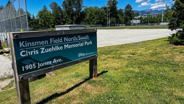 Teal Chris Zuehlke Memorial Park sign beside the Kinsmen sports field, an open gravel ground ringed by fencing and floodlights with mountains rising beyond.