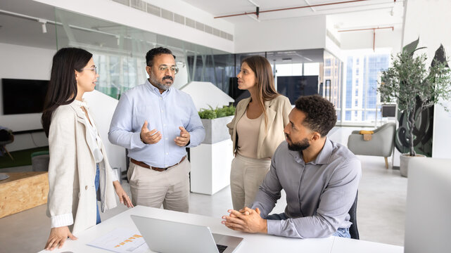 Group of professionals engaged in discussion in modern coworking office, listening opinion and ideas speaking Indian mature team leader, gathered together in contemporary corporate workspace. Briefing