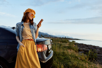 Fototapeta premium Cheerful woman outdoors, wearing striped top and yellow skirt, leaning against a car by the coast, enjoying a sunset moment, casual lifestyle, natural colors