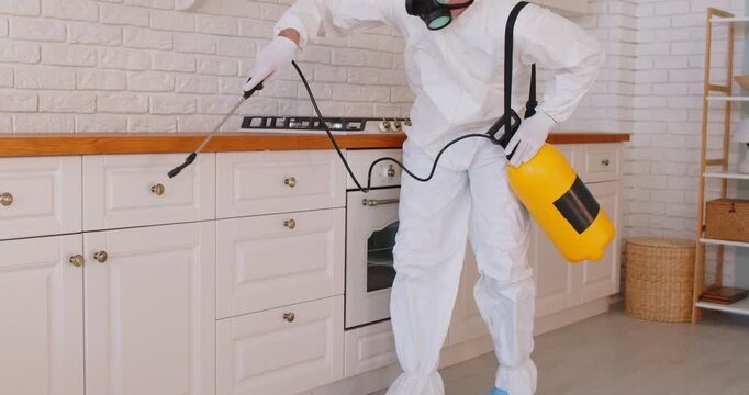 Exterminator with sprayer disinfecting modern kitchen furniture to eliminate cockroaches. Pest control worker in white protective suit spraying to apply insecticide for disinfection, kill insects