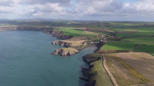 Aerial view of the rugged Welsh Coast featuring steep cliffs, lush green fields, and a small bay with boats under a cloudy sky in St Davids, Wales, United Kingdom.