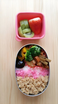 A Japanese homemade bento lunch box with minced chicken (soboro) and pink sakura denbu on rice, broccoli, kinpira gobo, simmered pumpkin, and a separate pink container with strawberries and kiwi fruit