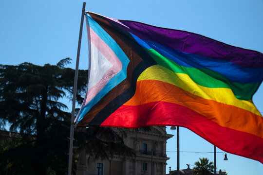 Bandiera del Progress Pride che sventola nel cielo azzurro durante una manifestazione, simbolo moderno di inclusione per la comunit&agrave; LGBT, persone transgender e minoranze etniche