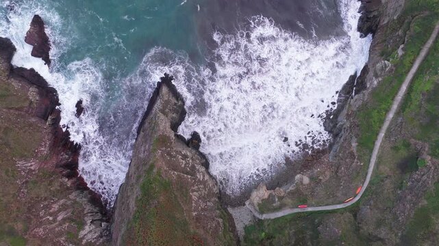 Aerial view of waves crashing against the rugged cliffs of the Welsh Coast with a narrow path winding along the green hills in St Davids, Wales, United Kingdom.