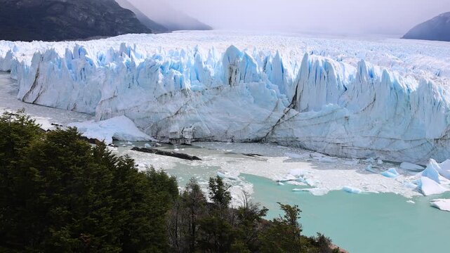 Aerial view of the massive Perito Moreno Glacier with blue ice walls and floating ice chunks near green forests in Perito Moreno, Santa Cruz Province, Argentina.