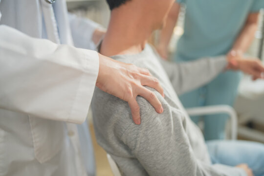 Asian nurse holding patient's hand while doctor examines shoulder and arm during physical therapy session, healthcare team assessing injury in clinic with bright window light, medical examination, cop