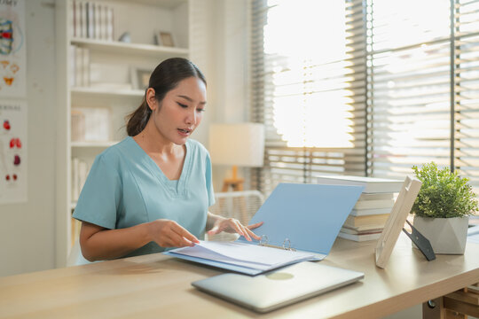Asian female nurse reviewing patient medical records in binder at desk, healthcare professional checking documents in bright clinic office with anatomy posters, medical administration concept, copy sp