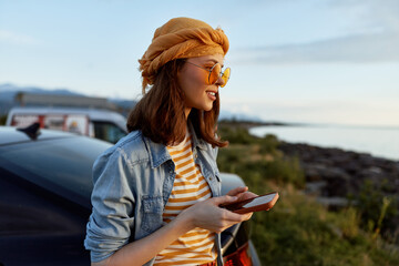 Fototapeta premium A stylish woman in a denim jacket and striped shirt checks her phone beside a car, enjoying a sunny outdoor coastal moment. Casual lifestyle concept.