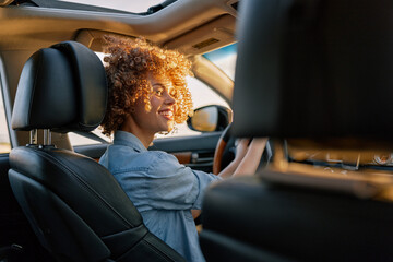 A smiling young woman with curly hair drives a car, wearing a denim shirt, sunlight on her face, capturing a candid moment of everyday mobility and independence.