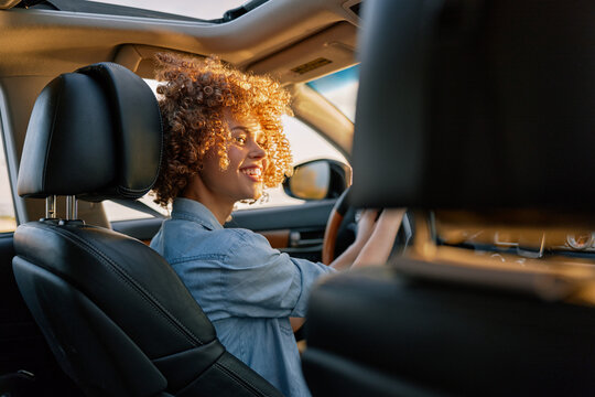 A smiling young woman with curly hair drives a car, wearing a denim shirt, sunlight on her face, capturing a candid moment of everyday mobility and independence.