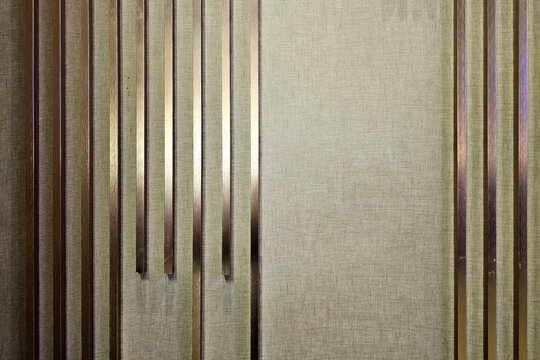 Modern Decorative Wall Panel Featuring Vertical Bronze-Colored Metal Louvers Against Textured Light-Colored Fabric Background, Contemporary Interior Design Element.