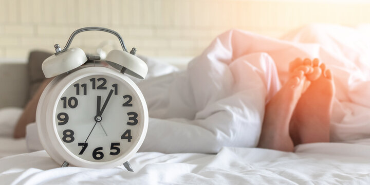 World sleep day, sleeping hour, health relaxation and bedtime insomnia concept with young girl&rsquo;s bare foot laying on bed resting in bedroom on white bed sheet with alarm clock