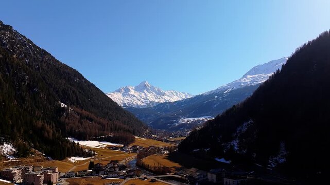 Aerial view of the alpine valley and town of Soelden surrounded by snow-capped mountain peaks and dense forests under a clear blue sky in Soelden, Tyrol, Austria.