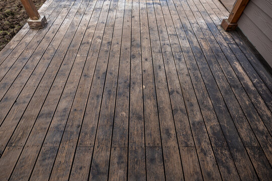 Weathered porch planks show gray black staining and patchy discoloration, framed by two square posts and house siding, daylight scene, low oblique angle, needs refinishing.