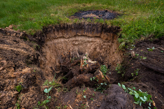 Large pit with exposed soil layers and jagged roots sits in short grass. Charred ground lies farther back. Daylight, overcast light, low angle, damp earth tones, parkland or farmland.