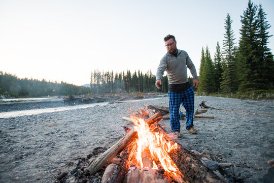 Man in plaid pajama pants tends campfire by river