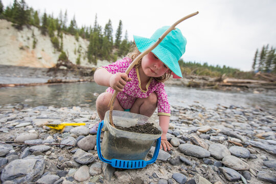 Toddler girl plays with a stick and bucket by the river