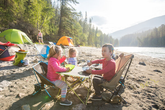 Three young kids eat breakfast at a riverside campsite