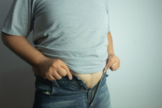 Close up of overweight man struggling to close tight jeans button over his big belly. Weight gain realization, abdominal obesity, and diet challenge concept.