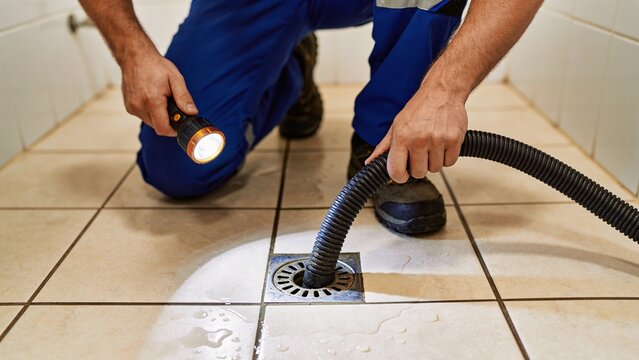A professional plumber using a flashlight and a vacuum hose to clean a clogged bathroom floor drain.