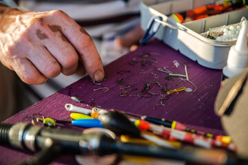 Fototapeta premium A person lays out fishing gear, mainly hooks, on a purple surface. Fishing floats, sinkers, a spinning rod, a fishing box and a tube of glue are visible. High quality photo