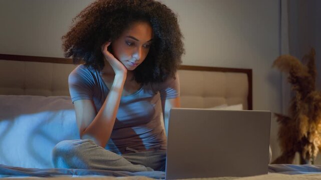 Sleepy boring African American woman looking at laptop screen in bed at night tired female girl using computer internet addiction browsing online cyberspace in dark bedroom insomnia sleep disorder