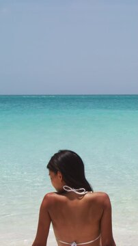 Vertical video. Beach vacation dream woman enjoying summer holiday on dreamy perfect ocean tropical destination. Person sitting from the back on white sand beach getaway. Panning up to Copy space.