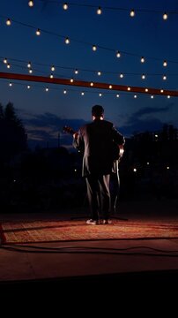 Man Speaking on Outdoor Stage at Dusk Under Hanging String Lights