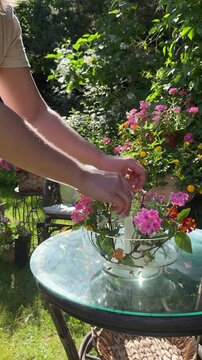 Woman places a white floating candle into a glass bowl with fresh flowers on a glass garden table. Cozy summer backyard with blooming flowers and natural sunlight. Vertical lifestyle video.