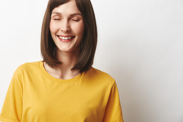 Fototapeta premium Young woman with shoulder length brunette hair wears a bright yellow tshirt and smiles softly while standing against a clean white wall, conveying a warm, relaxed mood and friendly approachable vibe