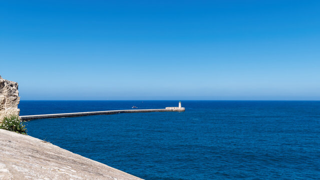 Long stone breakwater wall stretching into the deep blue sea towards a distant lighthouse, Valletta, Malta