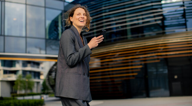Woman smiles while holding smartphone outside modern office building