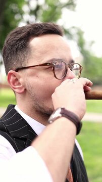 Man smoking cigar outdoors close up profile. Elegant gentleman with glasses and beard enjoying tobacco cigar in park, luxury lifestyle, retro fashion and wealth concept