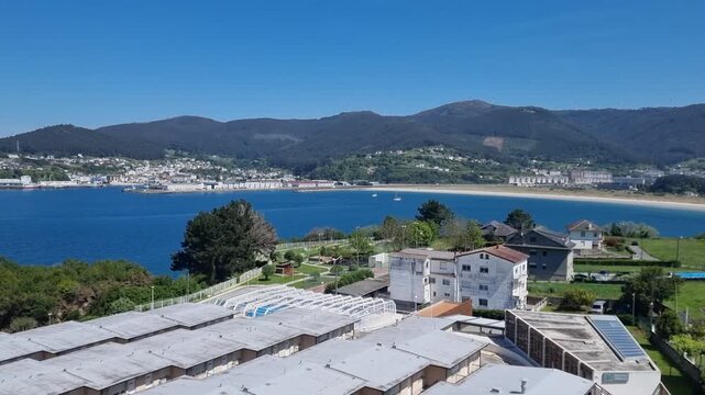 A sweeping panoramic view of Viveiro town curved around the sandy bay and blue sea of the Covas beach with rolling green mountains under a clear sky in Galicia, Spain.