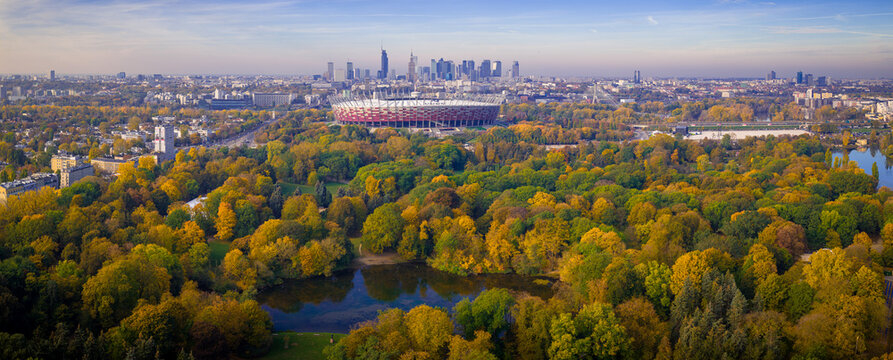 Warszawa, Poland - 22 October 2024: Aerial view of PGE Narodowy stadium surrounded by autumn trees in Skaryszewski Park with the city skyline in the background.