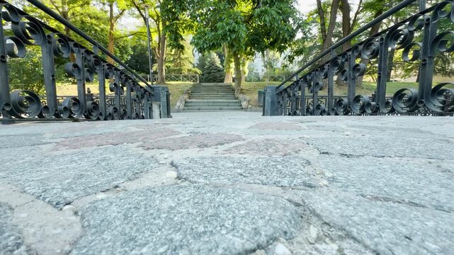 A stone bridge path opens up to a lush green park in Simferopol, Crimea. The serene walkway creates a peaceful atmosphere. Sunlight warms the vibrant trees ahead.