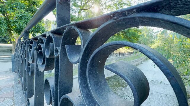 An ornate black iron railing frames a calm flowing river in a peaceful park. Tiny midges swarm gently in the bright morning sun, creating a serene and wondrous feeling.