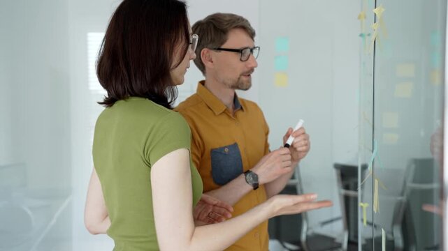 Business colleagues collaborate at a modern office glass board, mapping a timeline with sticky notes and hand drawn charts during an engaged strategic planning session