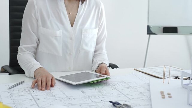 Woman architect analyzing blueprints and architectural models with a digital tablet on a clean desk, working on a construction project in a bright office environment
