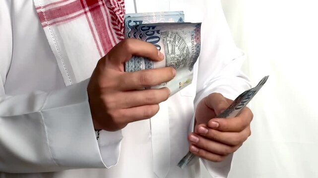 Saudi man wearing traditional thobe counting five hundred riyal banknotes close up.