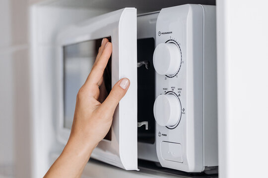 Hand of a person opening a white microwave oven door with control knobs visible, showcasing a modern kitchen appliance in a bright interior setting