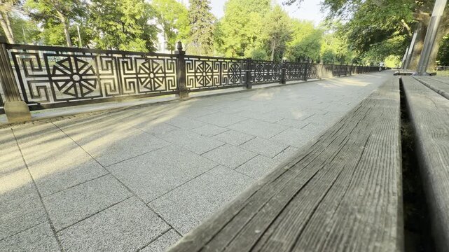 A clean stone walkway rests quietly beside a beautiful decorative fence. The sunny park path feels calm, inviting, and peaceful under green trees.