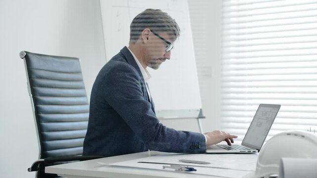 Architect or engineer working on construction blueprints and plans at an office desk, using a laptop for design and planning, embodying professionalism and project management