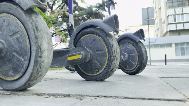 Electric scooters stand parked on a paved city sidewalk. Their dusty wheels rest quietly, evoking a calm and lonely feeling.
