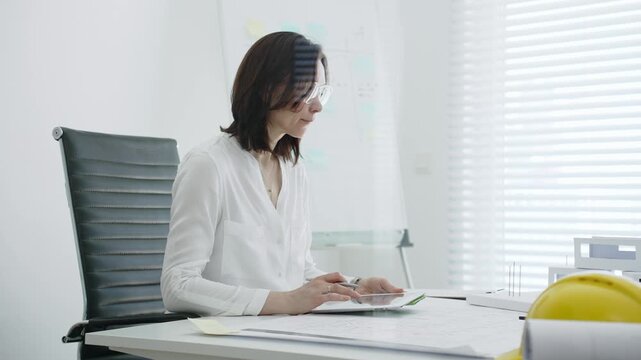 Female architect analyzing blueprints and architectural models in office, using digital tablet for modern design planning and engineering project development